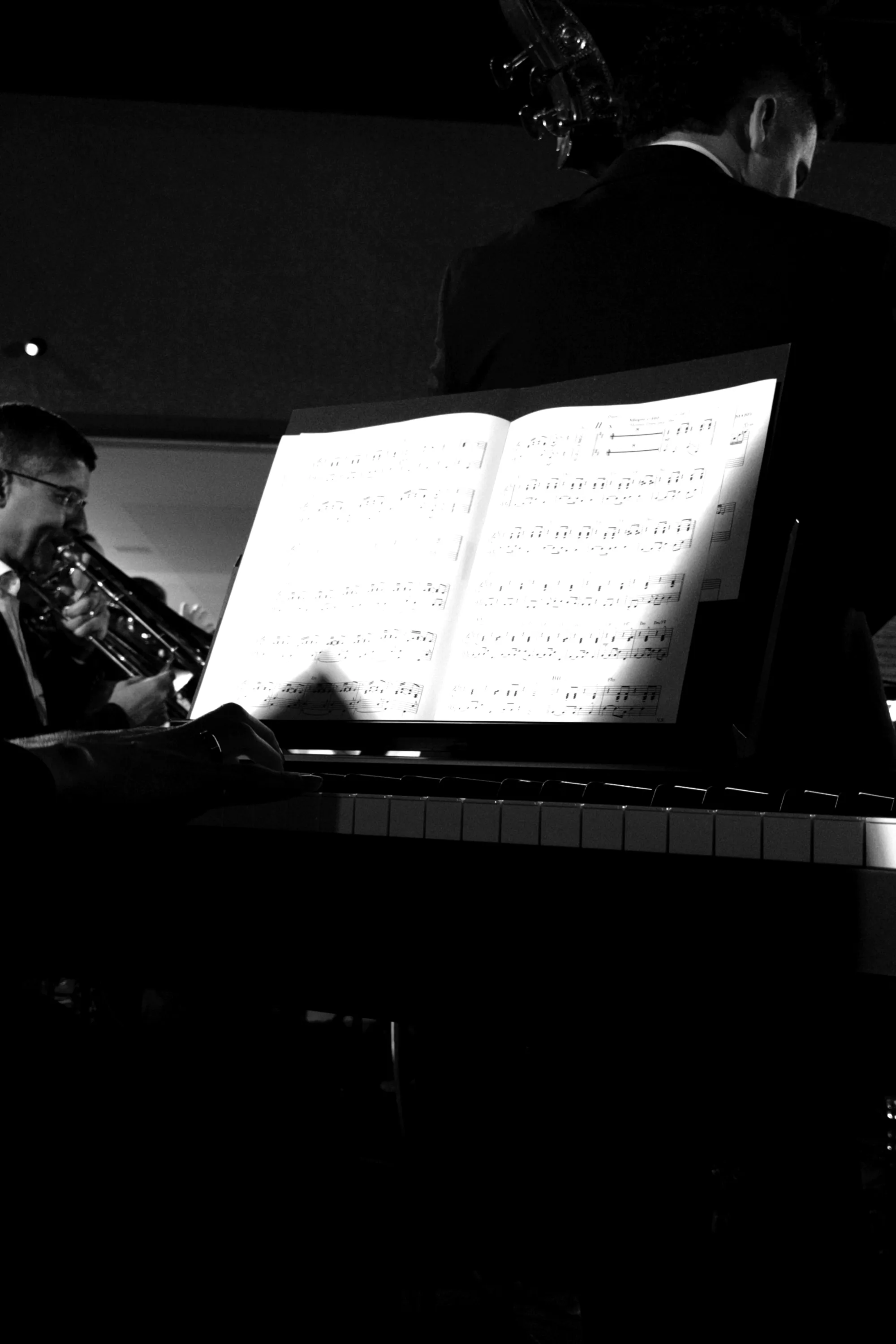 Partitura iluminada no estande, músico de trombone ao fundo, Filarmônica Aruna em ensaio