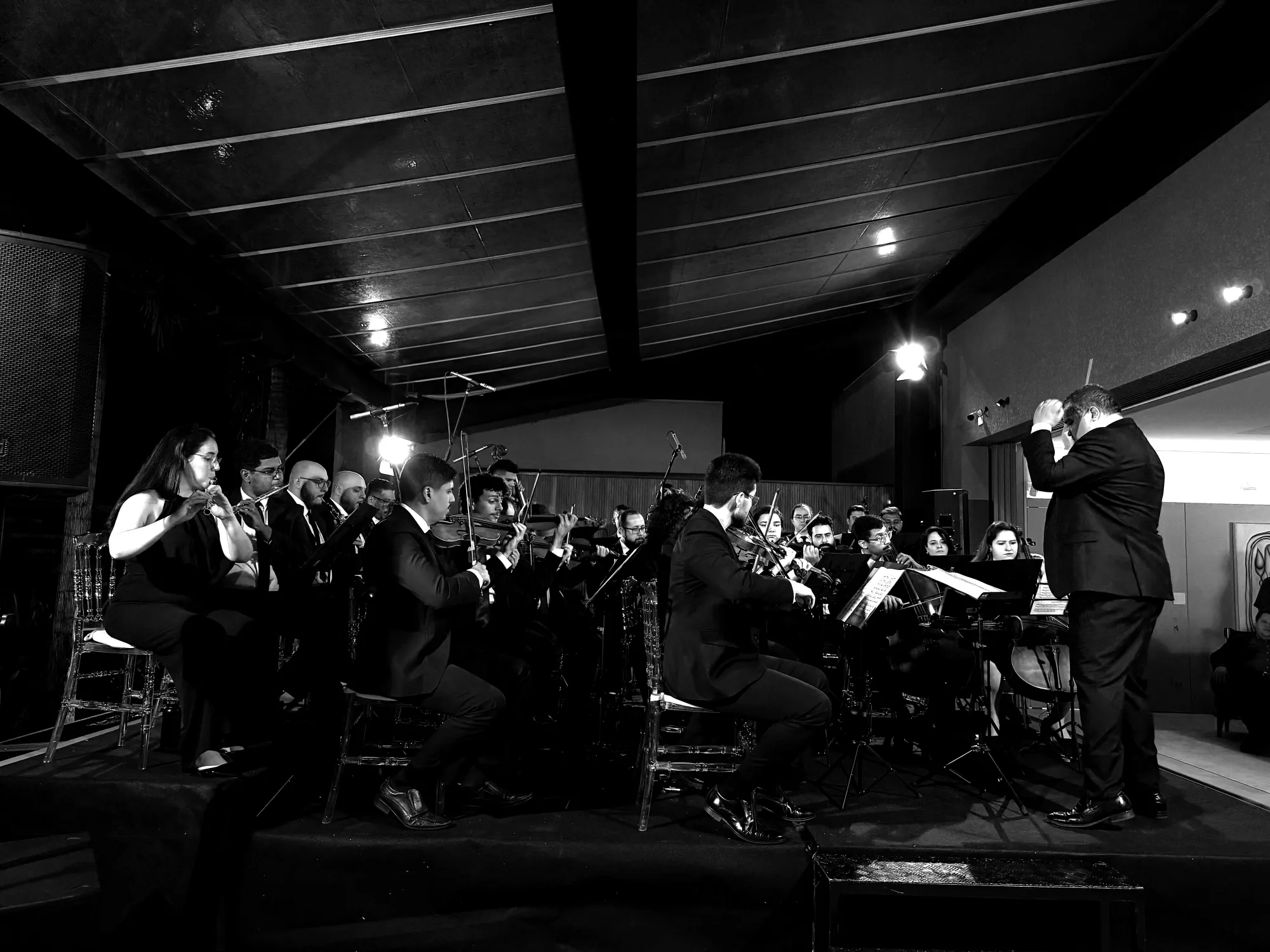 Filarmônica Aruna em apresentação, maestro regendo à direita, orquestra completa ao fundo