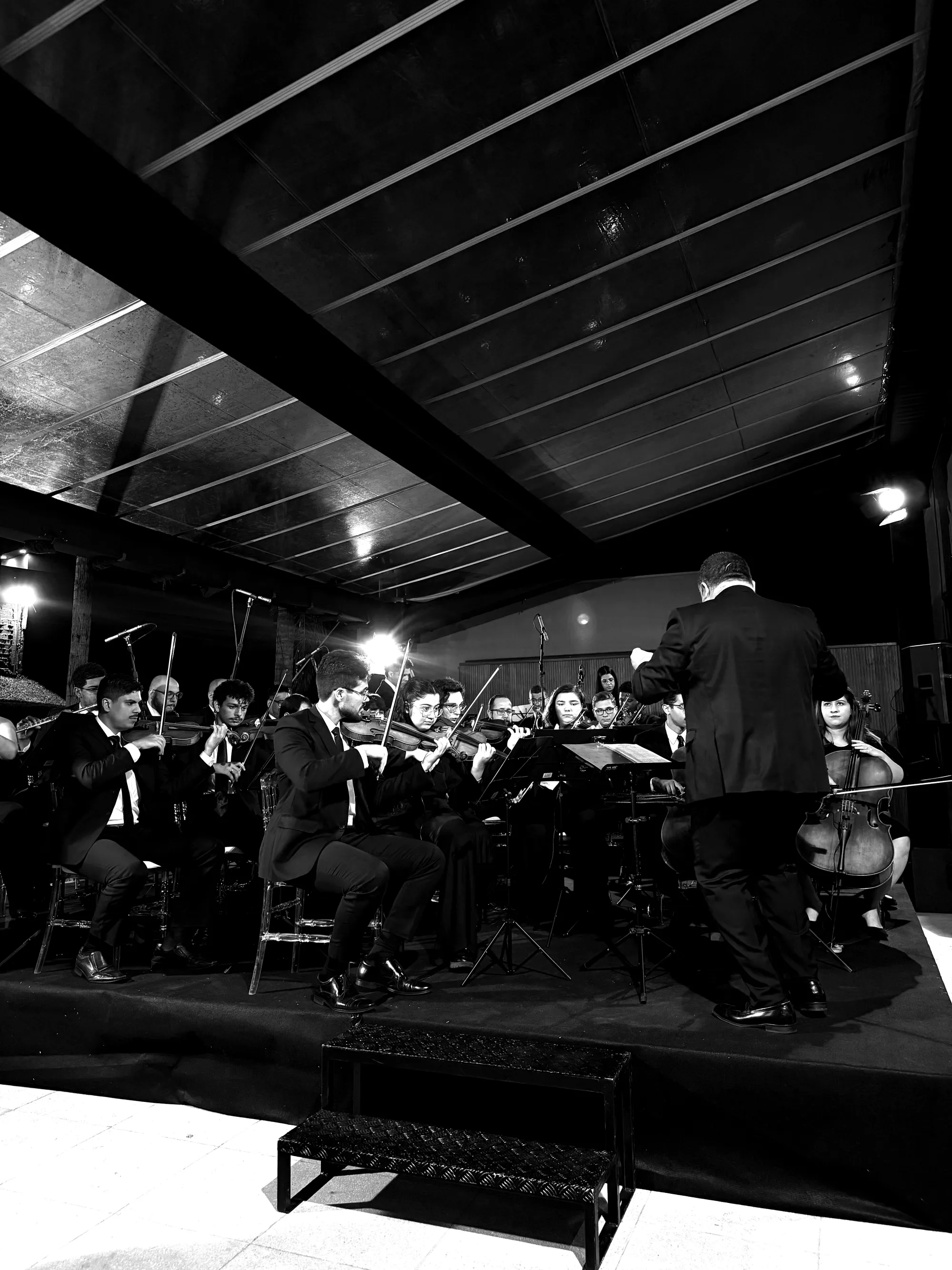 Filarmônica Aruna completa em apresentação, maestro regendo de costas, visão ampla do palco
